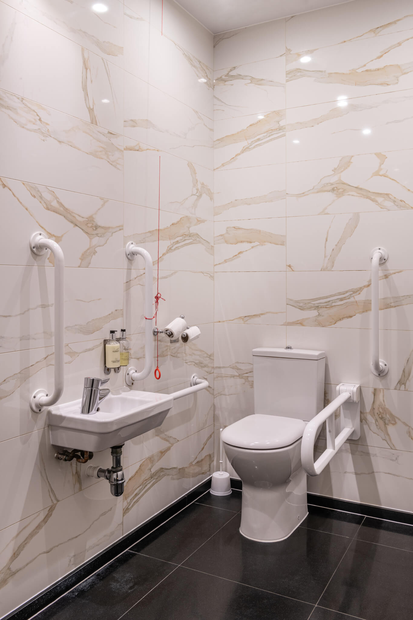 Clean, modern disabled toilet with matt white accessible bathroom equipment.  Beige and white marbled tiles.