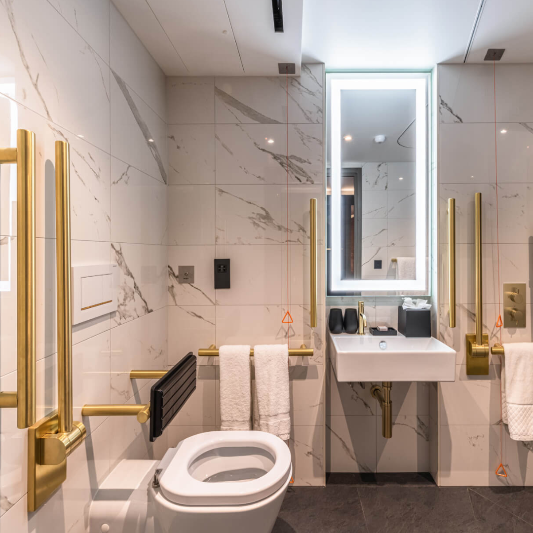 Elegant accessible bathroom with gold finishes on grab rails, hinged support rail, taps, and a black backrest above the toilet.