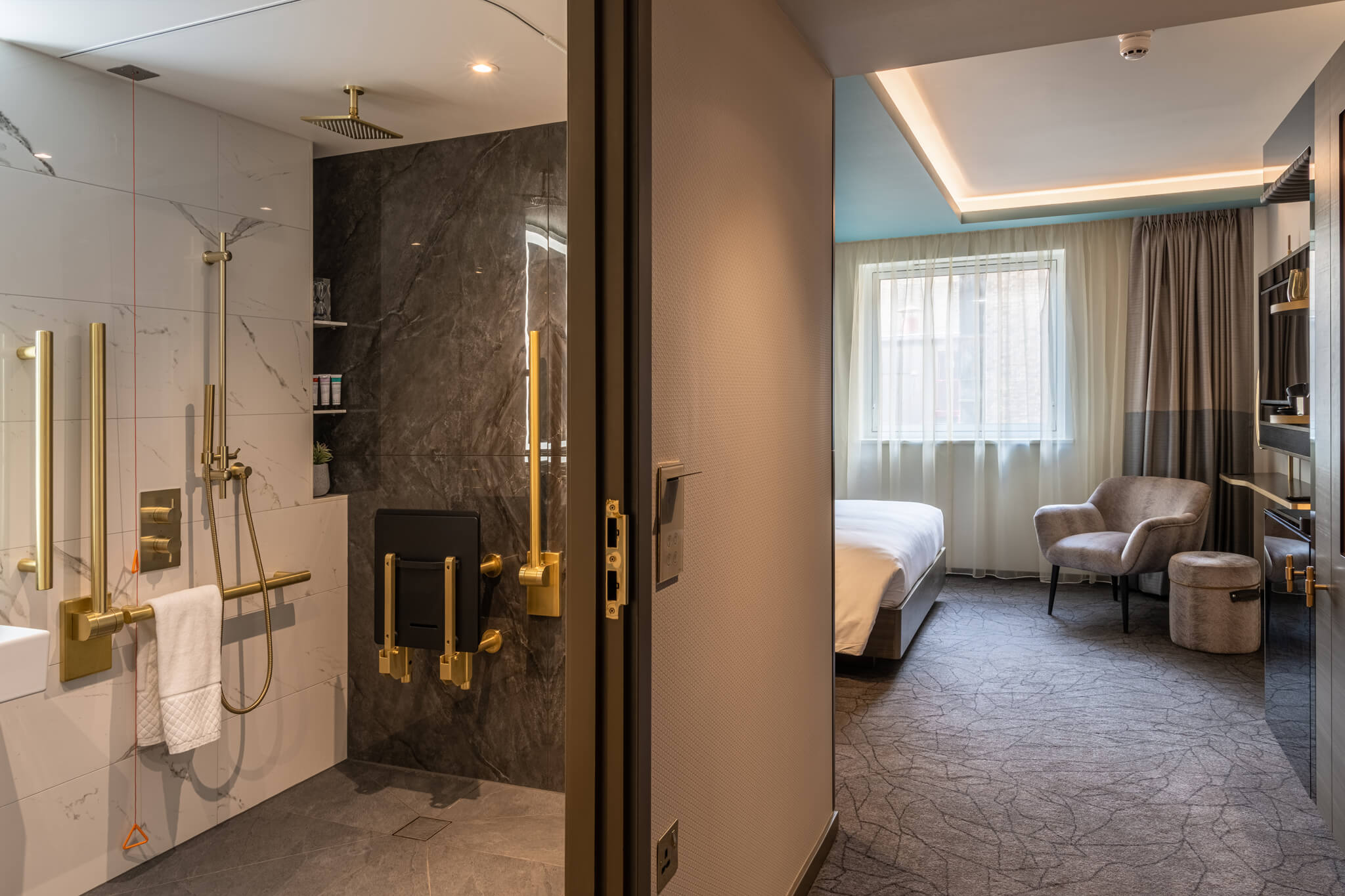 A luxury hotel room showing its accessible bathroom with shower. Finishes are all in gold, with white marble tiles on the walls. 