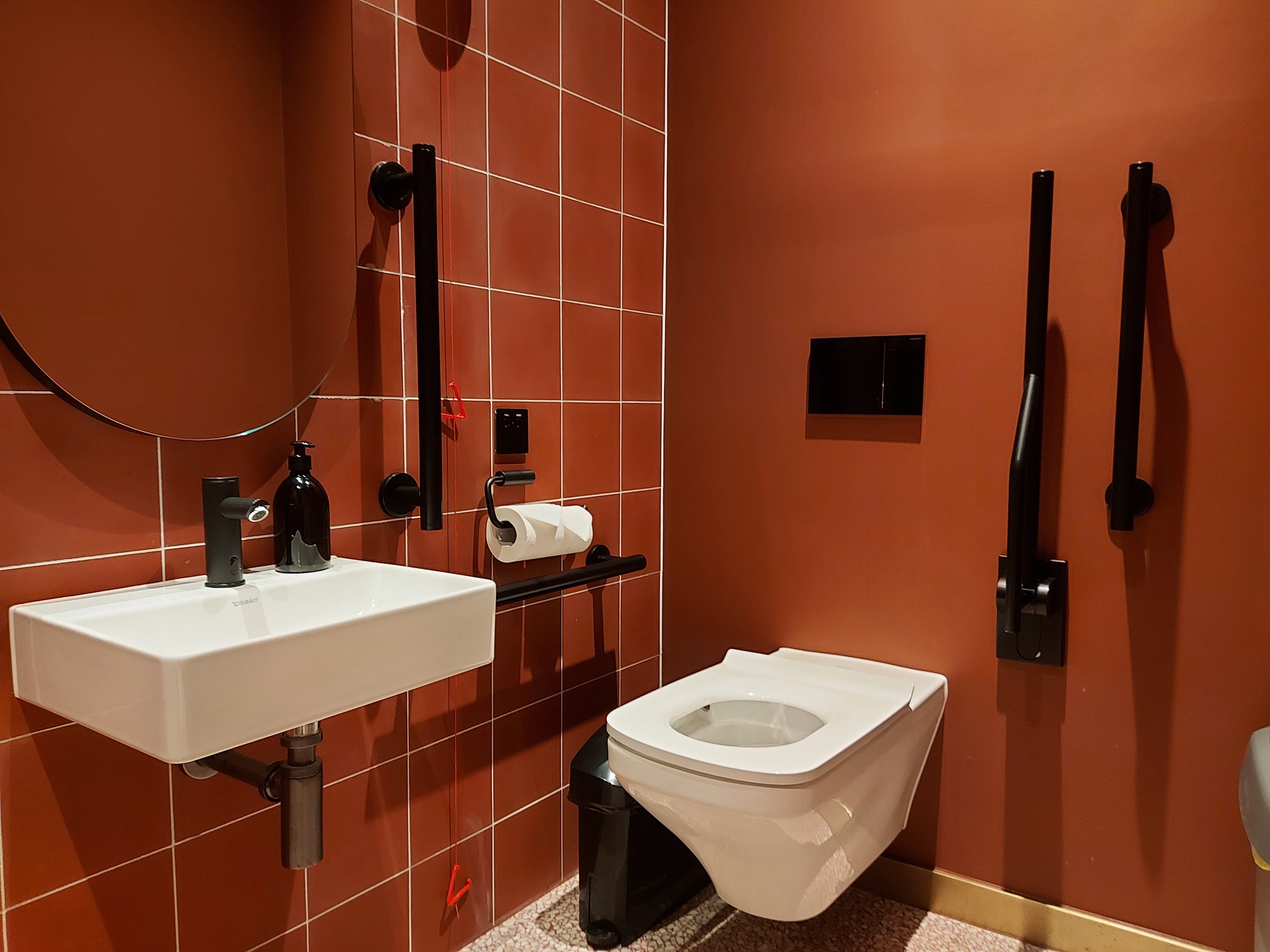 Guest toilet with orange tiles and walls, coupled with black, glossy grab rails and a hinged support rail folded up.