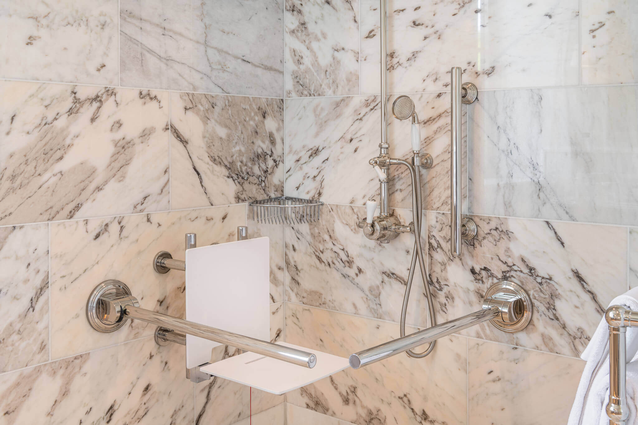 A accessible shower with removable showering seat in white and stainless steel bathroom taps, grab rail and support rail. Shower wall tiles are marbled in white and grey. 