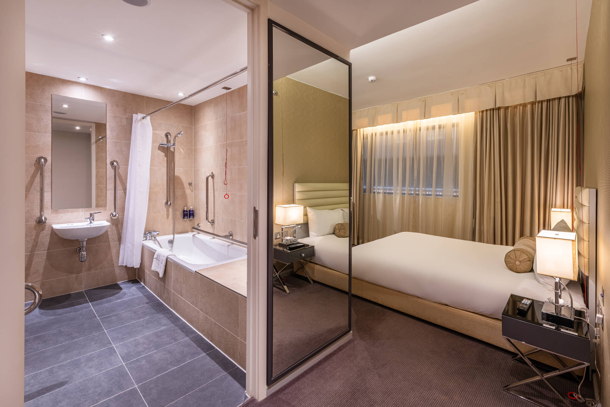 Stylish hotel bedroom and bathroom that has stainless steel showering taps and horizontal grab rails. 