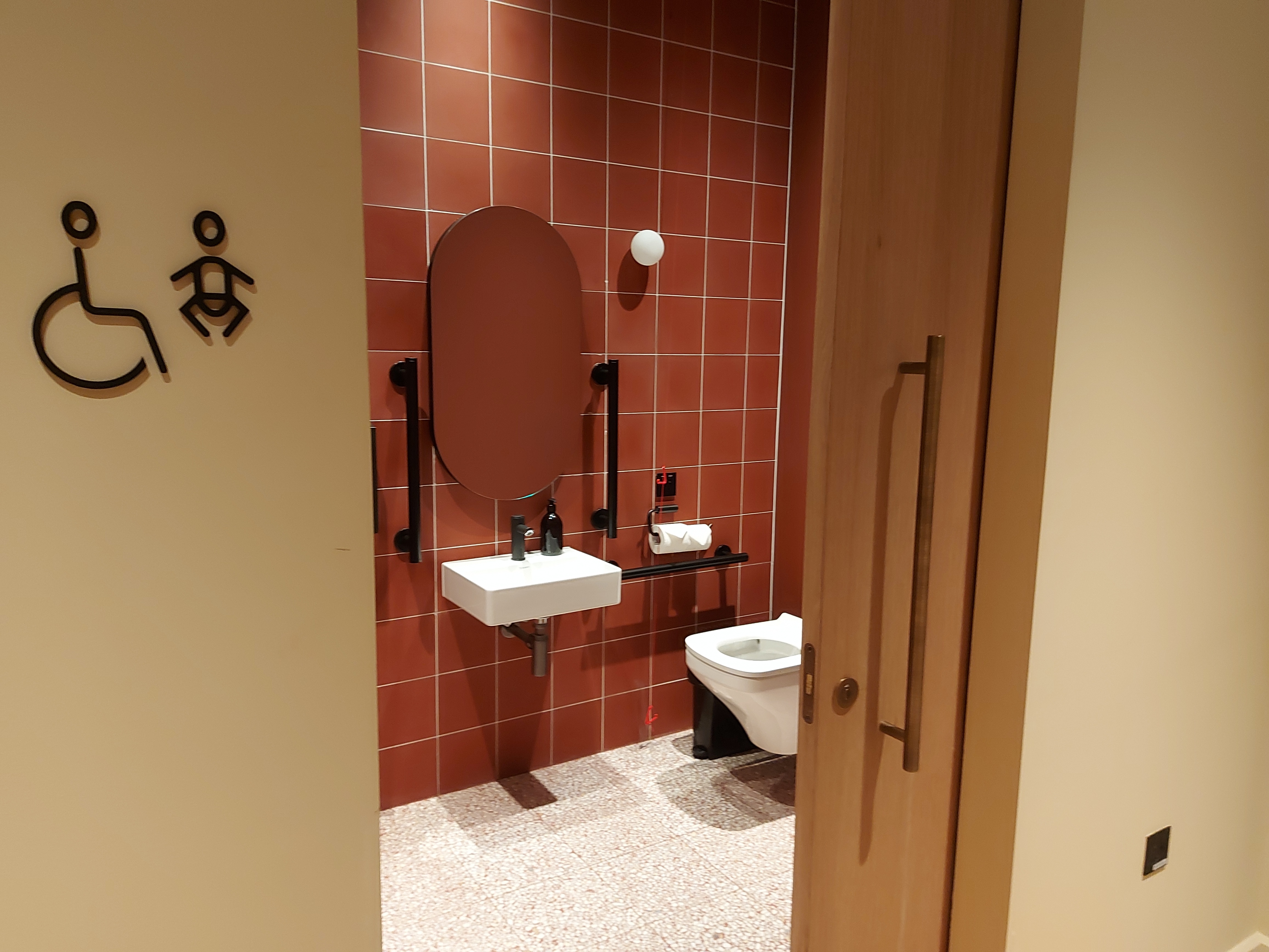 A public, inclusive toilet decorated with dark orange tiles and matt black grab rails by toilet and wash basin